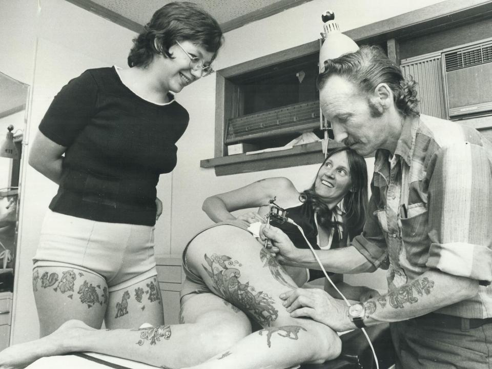 A woman smiles from a table as her husband shows how he applies tattoo.