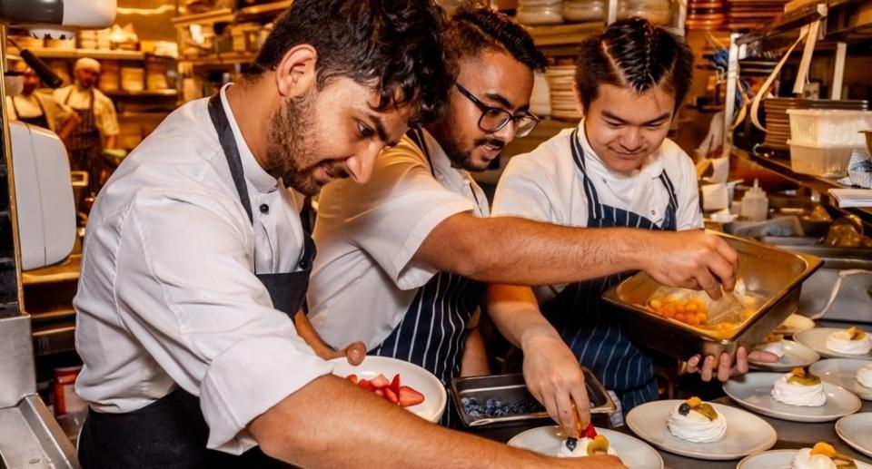 Three chefs enjoying preparing deserts in a busy kitchen .