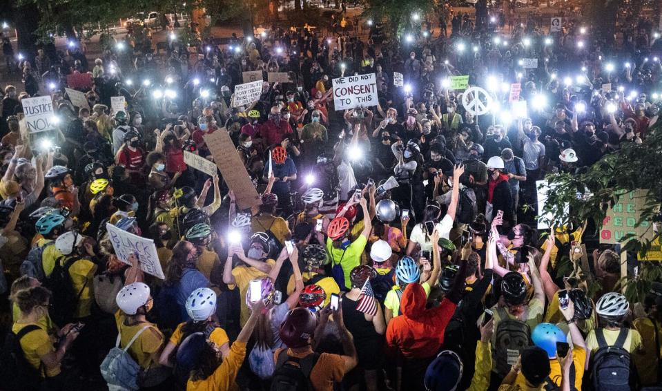 Protesters in bike helmets hold up signs and illuminated cellphones at a nighttime rally