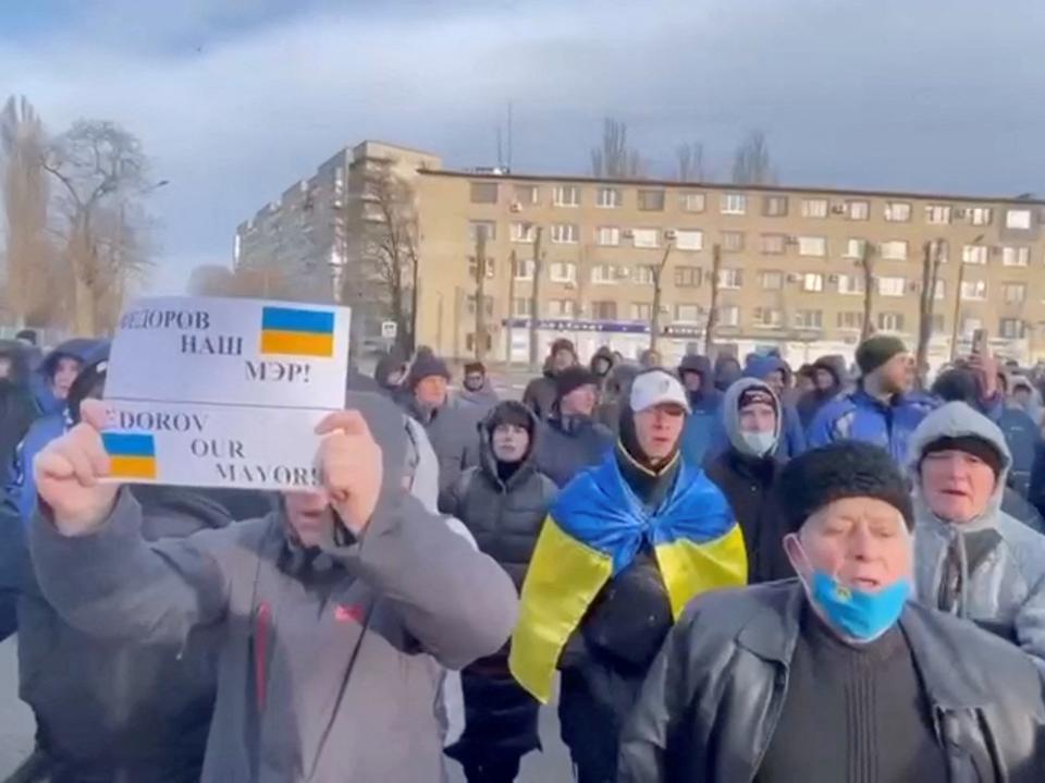 A crowd of Ukrainian men stand outside in Melitopol holding up signs in protest of the abduction of their mayor.