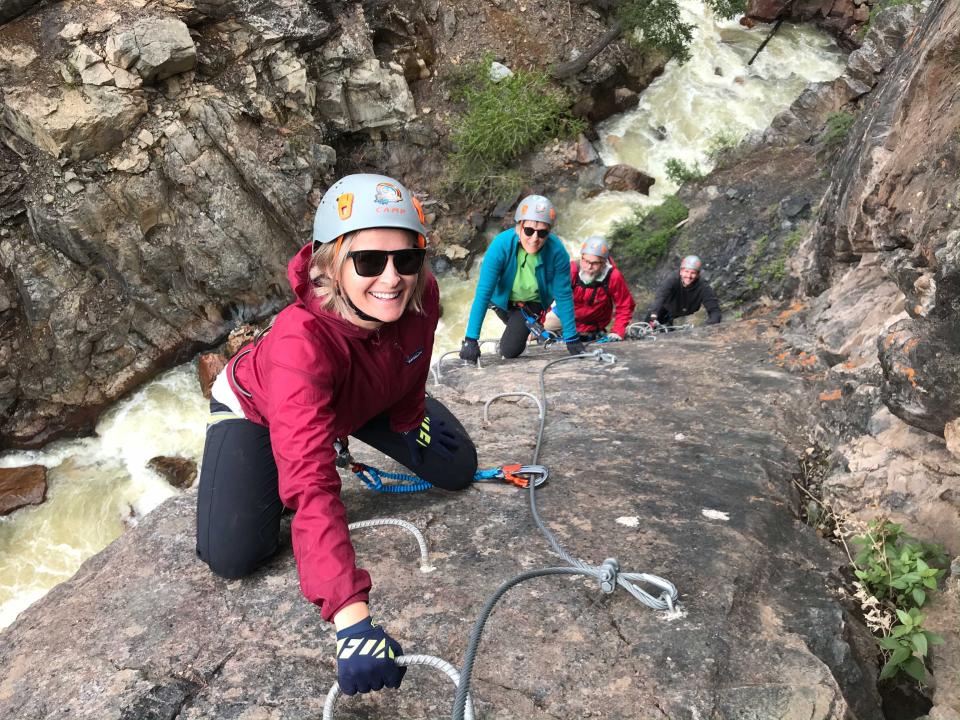 A woman rock climbing and smiling at the camera with a few people behind her.