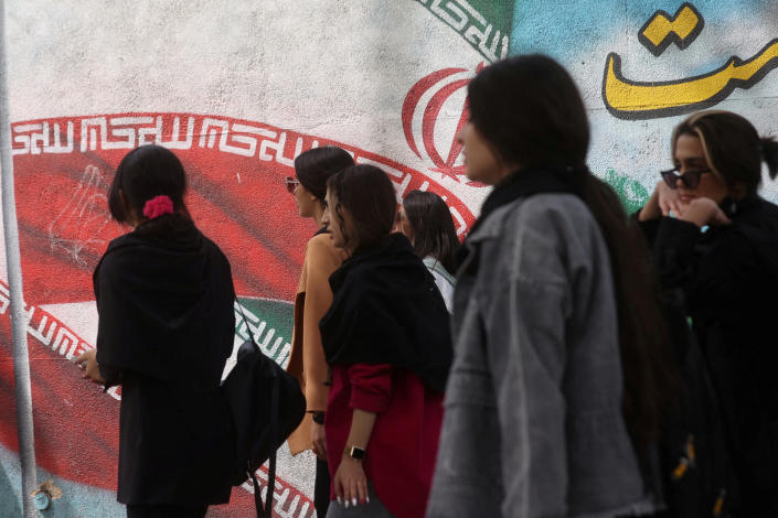 A group of Iranian women in suits and without the veil, one with bare arms, walk past a giant mural of the Revolution. 