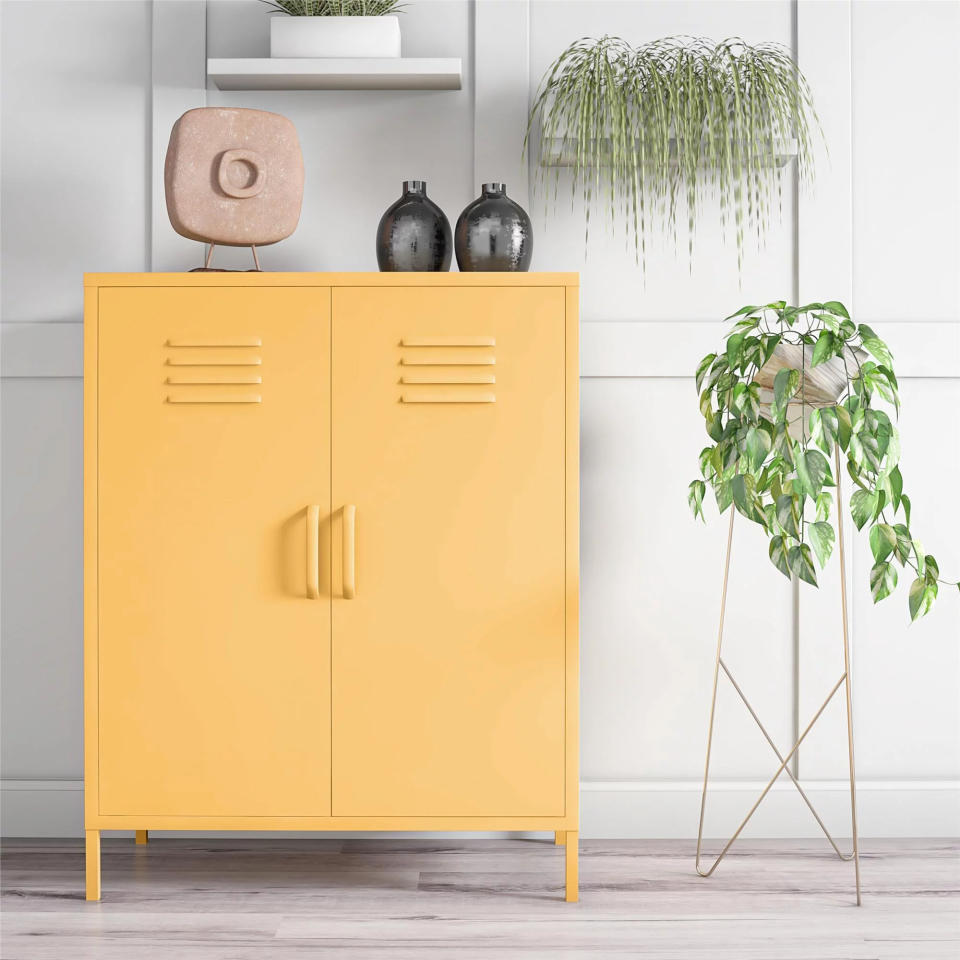 Yellow cabinet with dual handles, decorative vases, and a stone sculpture on top. Tall potted plant on a stand and indoor hanging plant nearby