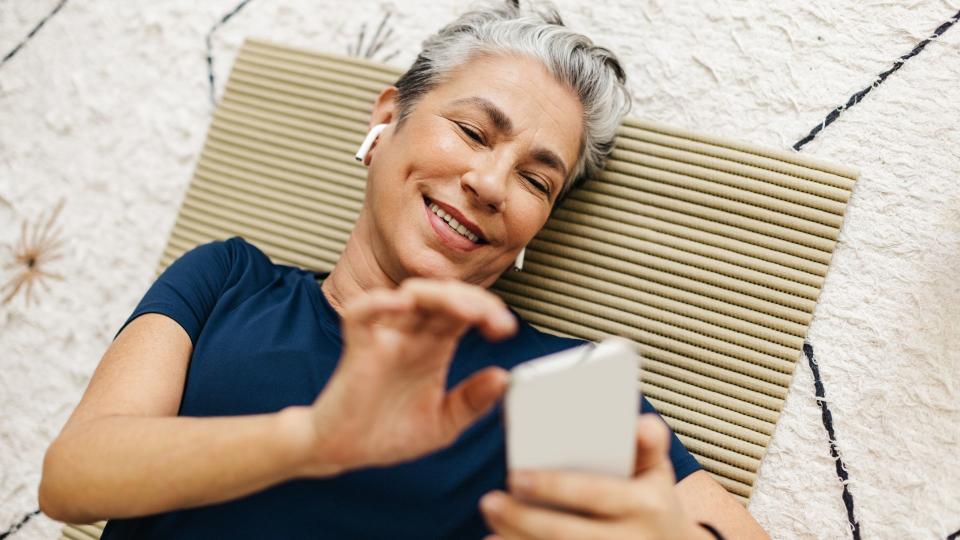 Woman lying back on mat with headphones and holding phone above face, smiling