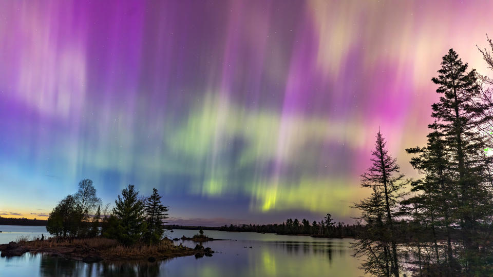     Nordlichter blitzen über einem See in Minnesota in einem dunklen Himmel über uns auf und erstrahlen in einem Regenbogen aus Aurora Borealis-Licht, während die Sonne hinter dem Horizont untergeht. 