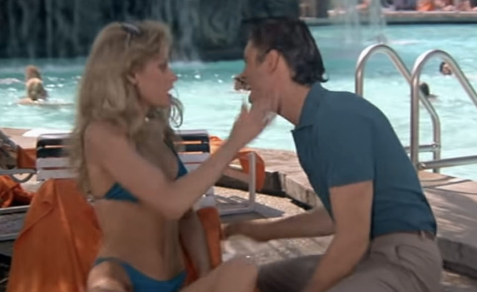 A woman in a bikini and a man in casual wear sit by a pool with waterfalls in the background, engaged in conversation