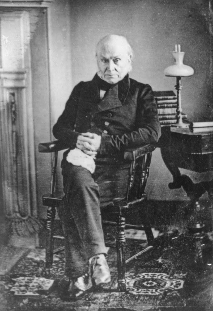 A balding, stone-faced John Quincy Adams sitting in a wooden chair with his legs crossed and his hands clasped