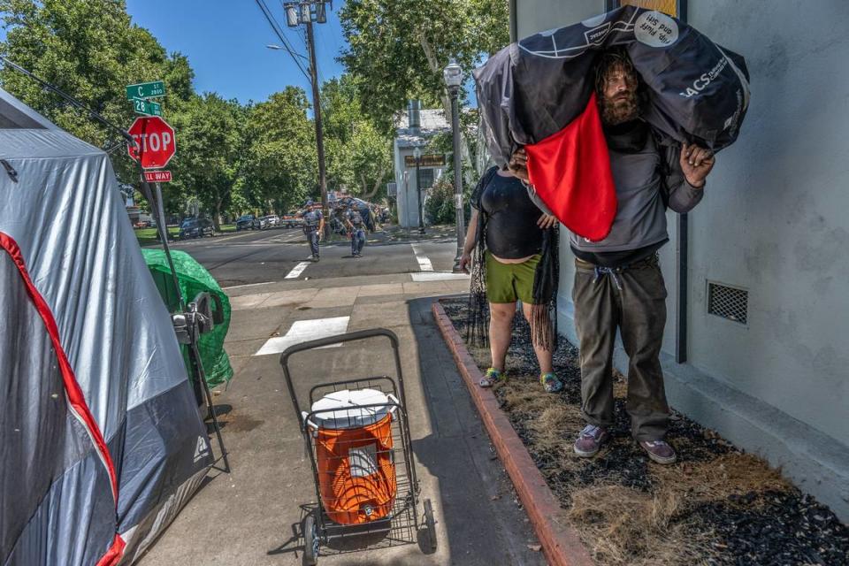 Homeless camper Chadwick Justin Foy, 41, holds all his belongings on his shoulders as friend Valencia Hardwich, 35, steps behind him in the shade while Sacramento police patrol the intersection of 28th and C streets on Tuesday, July 18, 2023. The city has given notices to dozens of people living in tents on the sidewalks in that area, near Leland Stanford Park, telling them to move by Wednesday. The notices cite violation of city codes regarding sidewalk obstruction and storage of personal property on public property. Hardwick said the church has been very supportive offering them water and food every week and the area was great because there were bathrooms in the park. They city has offered them Miller Park but she was concerned because it made people have curfews and said her friends relocated there said it’s a horrible mix of people that don’t get along. “People are a community here and we ban together where we are safe,” she said.