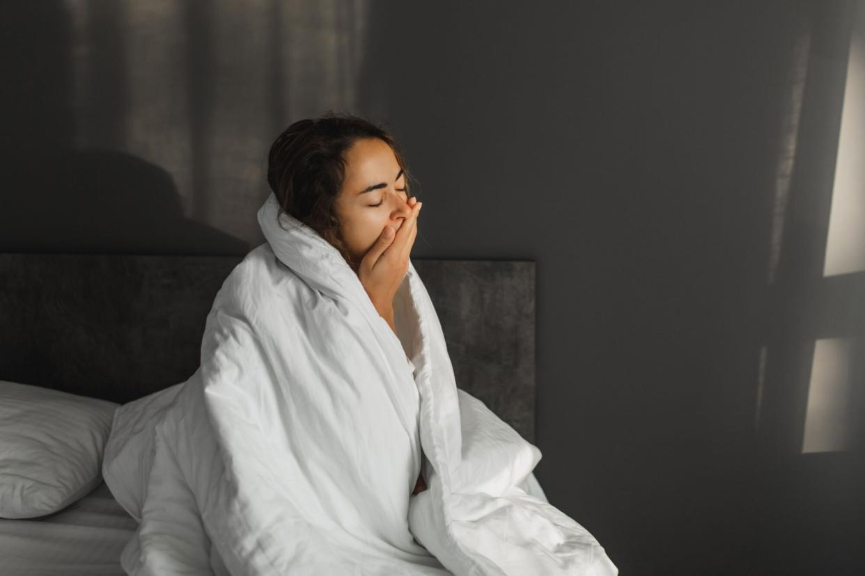 Woman yawning and can't wake up in morning. Sitting in bed covered with blanket.