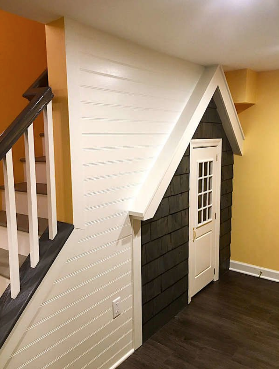 angular view of children's small house facade underneath a staircase in a basement with yellow walls