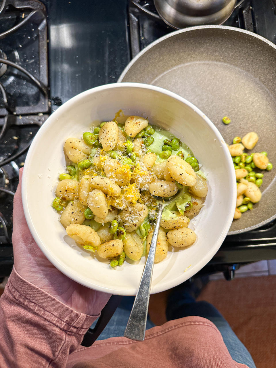 Hand holding a bowl of gnocchi with green peas, topped with grated cheese and lemon zest, in front of a stove and frying pan