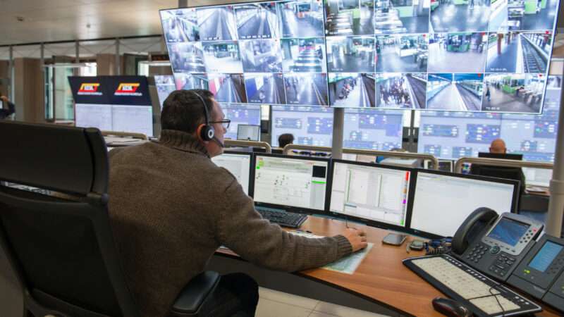 A man in an olive sweater sits in front of a bank of surveillance monitors.