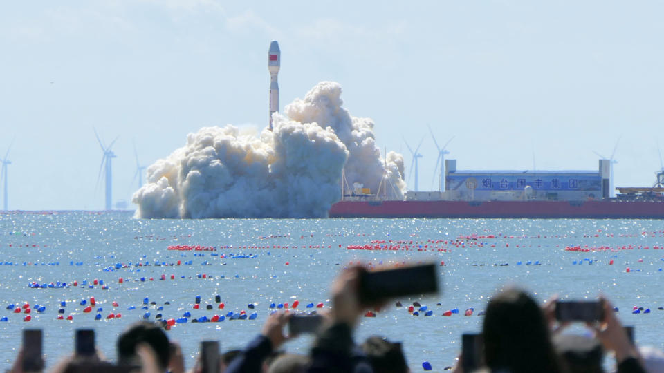  Spectators watch a white rocket launch into a blue sky from a ship anchored at sea, not far offshore. 