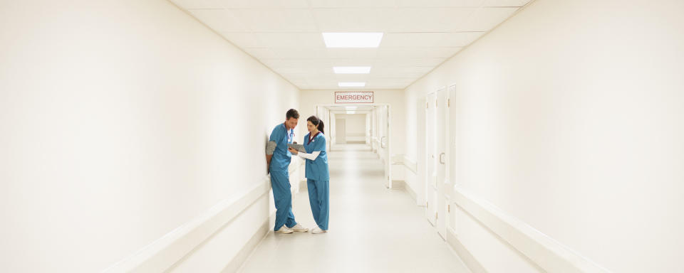 Two medical professionals in blue scrubs stand in a hospital hallway, engaged in conversation near an "Emergency" sign at the hallway's end