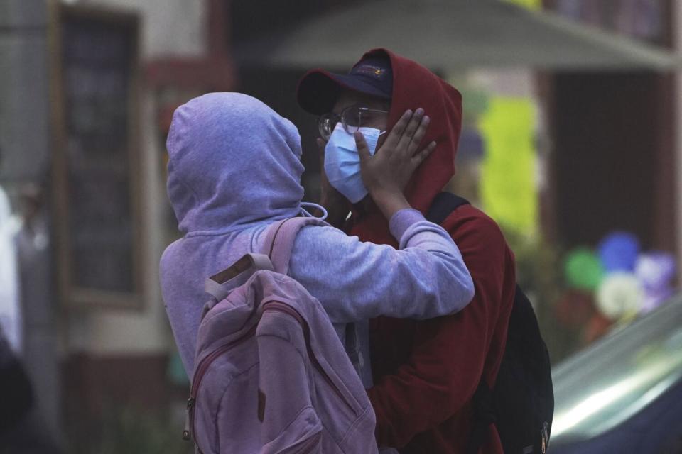 A person in a lavender hoodie and pink backpack places hands on the masked face of a person in a red hoodie, wearing glasses