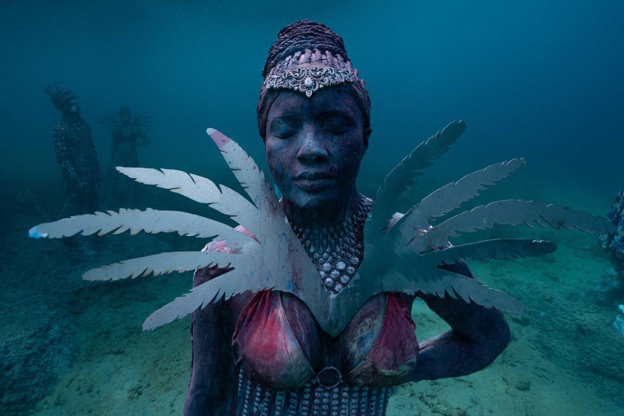 An underwater sculpture of a woman with her eyes closed, wearing an ornate headpiece, feathers, and an outfit splattered in red.