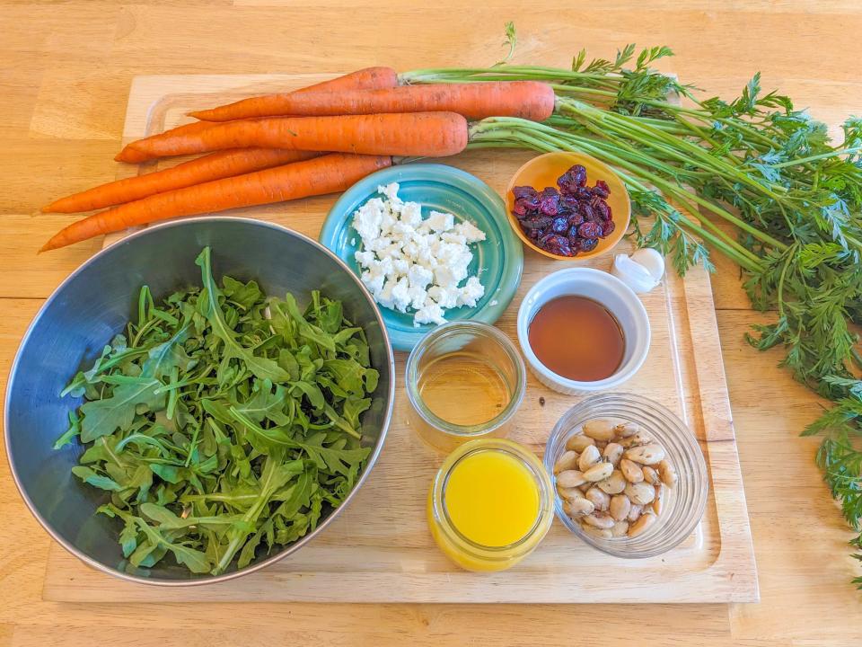 A wooden cutting board with bowls of carrot-arugula salad ingredients. The cutting board holds bowls of arugula, almonds, orange juice, cranberries, and cheese, as well as several carrots with the stems attached