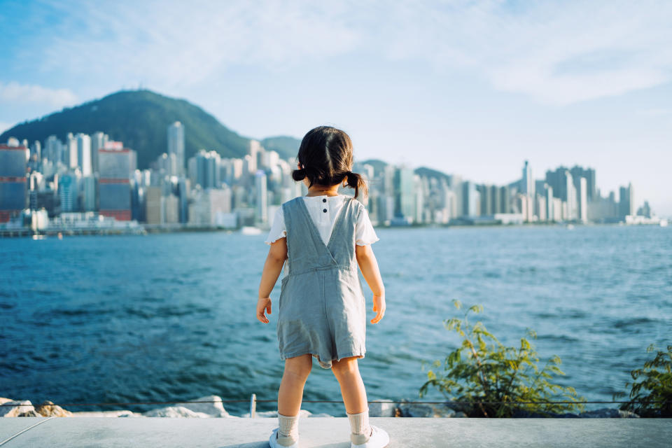 Rear view of cute little Asian girl enjoying the sun by the promenade and looking over city skyline and Victoria harbour on a lovely sunny day