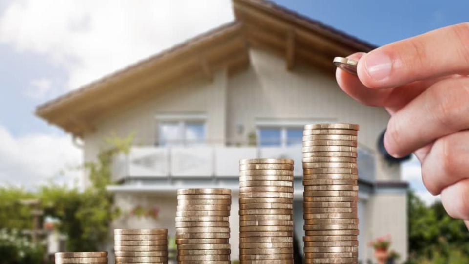 Human Hand Placing A Coin On Increasing Coin Stacks In Front Of House