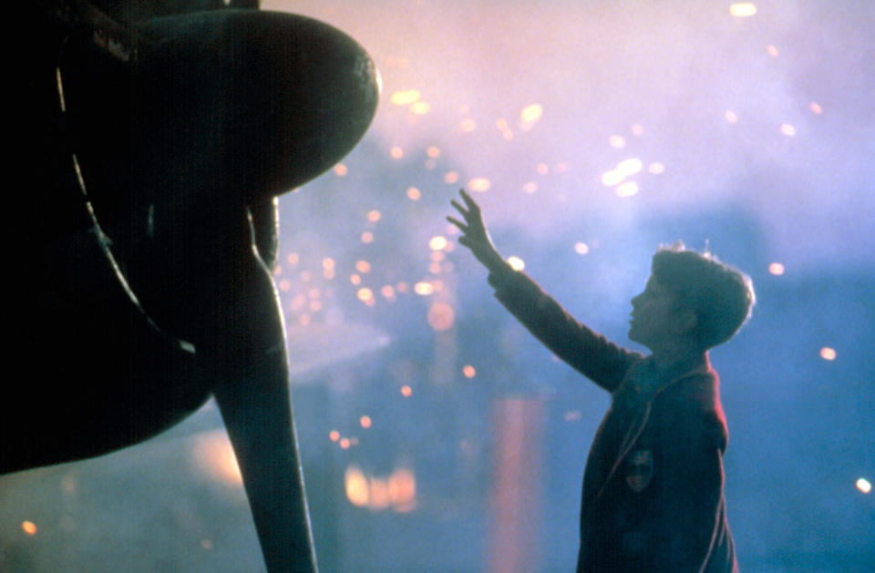 A young boy reaches up to touch the propeller of a plane as sparks fly in the background