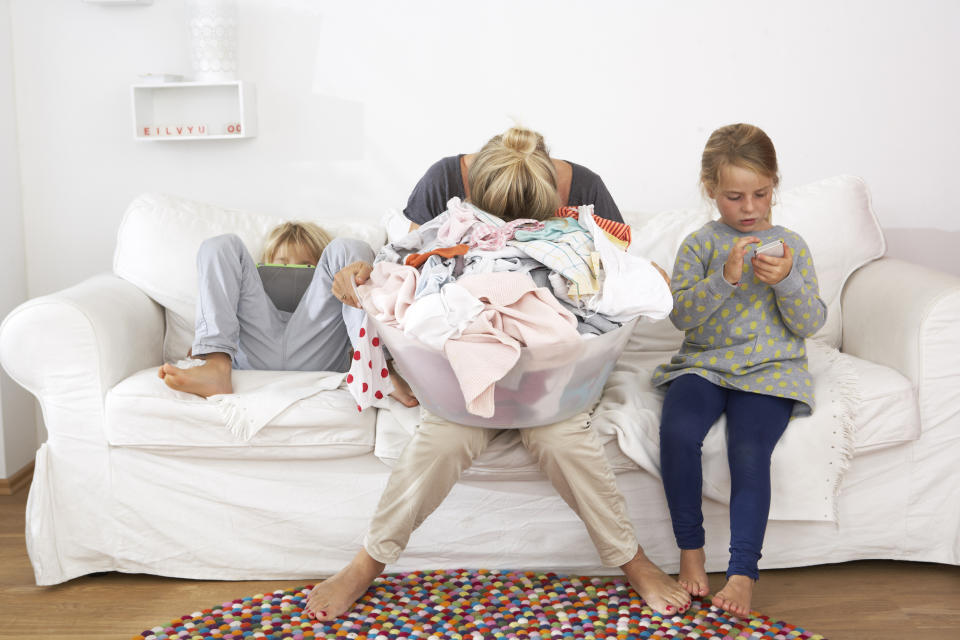 Exhausted mother with laundry basket on couch with children using digital tablet and cell phone