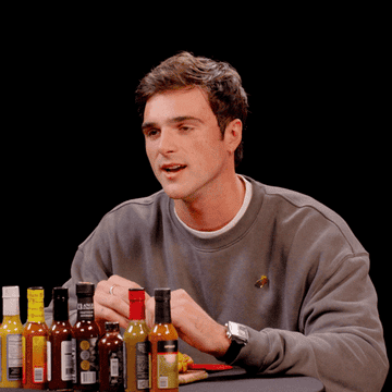 Jacob Elordi, wearing a casual crew-neck sweatshirt, sits at a table with various hot sauces in front of him. He says "hell no, man, absolutely not me, man"