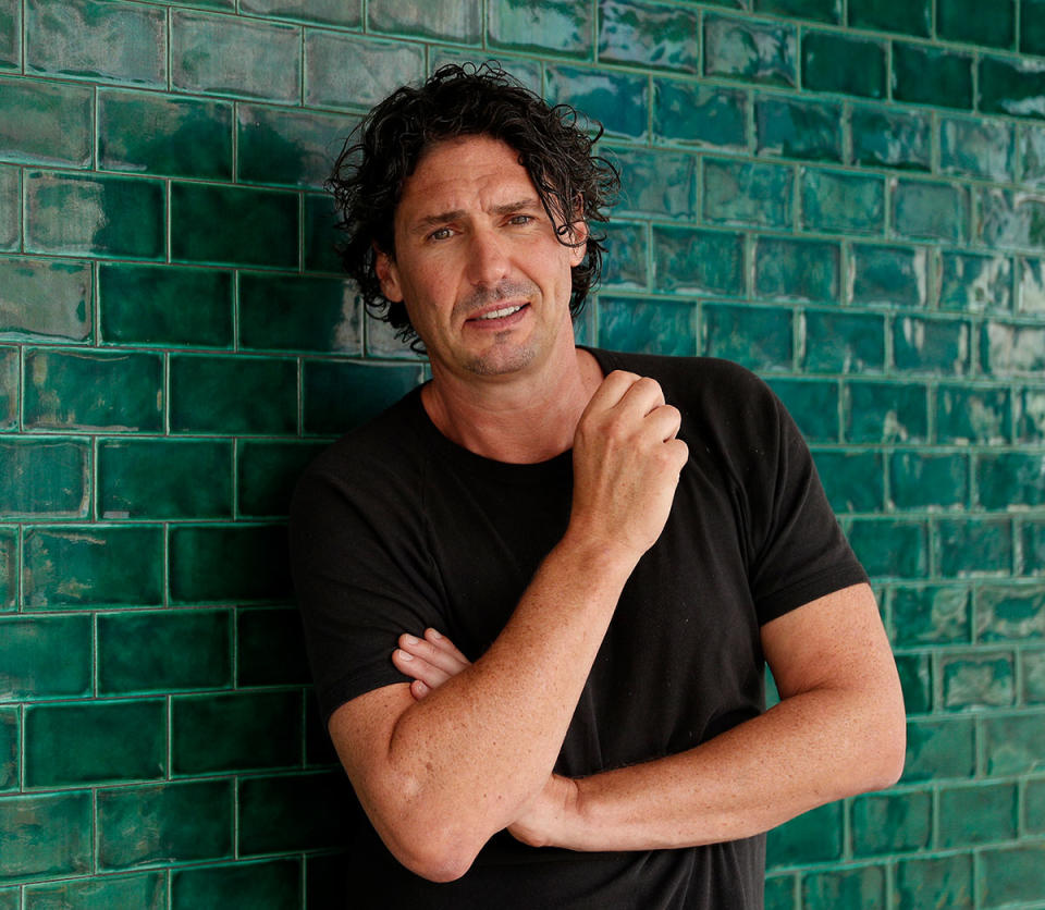 Colin Fassnidge wearing a black T-shirt standing against a brick wall