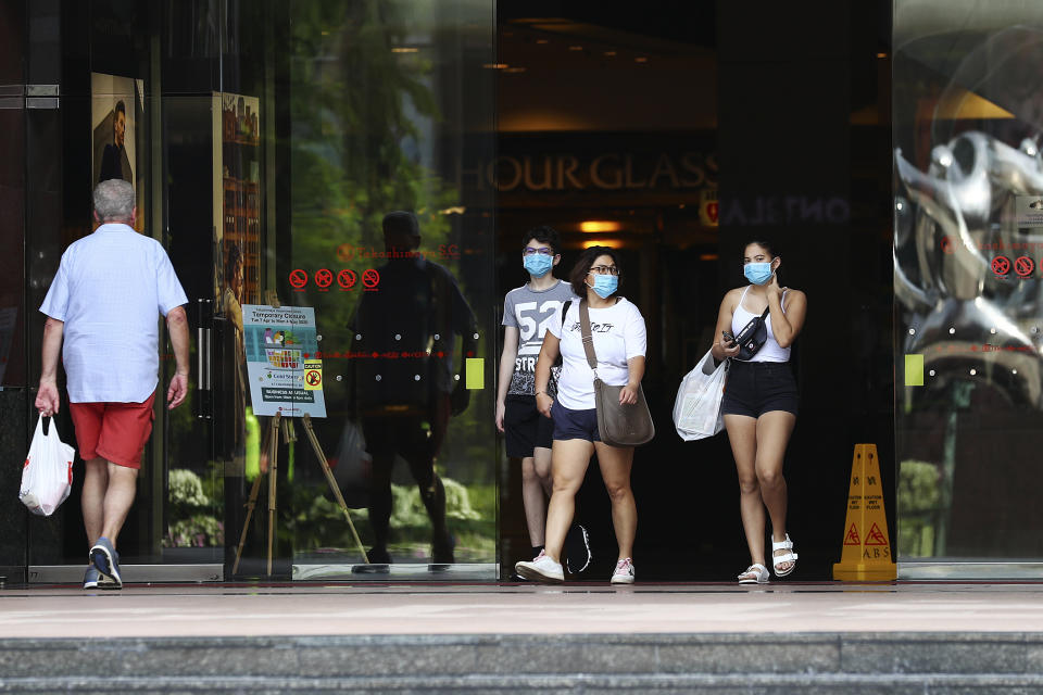 FILE - In this April 10, 2020, file photo, people wearing protective face masks exit a mall along the Orchard Road shopping belt in Singapore. A second wave of coronavirus infections in tightly packed foreign workers' dormitories has caught Singapore off guard, and exposed the danger of overlooking marginal groups in a health crisis. Infections in Singapore, an affluent Southeast Asian city-state of fewer than 6 million people, have jumped more than a hundredfold in two months — from 226 in mid-March to over 23,000, the most in Asia after China and India.(AP Photo/Yong Teck Lim, File)