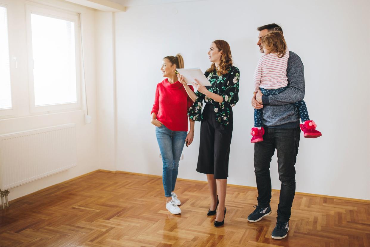 Real estate agent showing a room of a house to family, man is holding female child, room has white walls with two windows and wooden floors