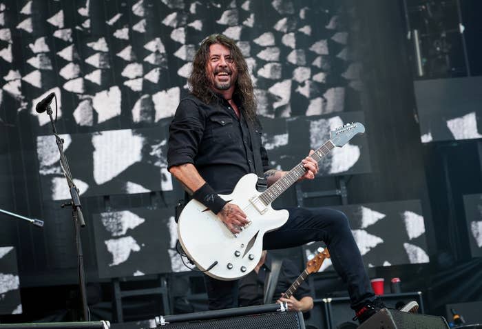 Dave Grohl on stage, playing a white electric guitar, smiling and dressed in a black outfit, in front of a geometric pattern background