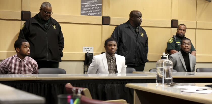 Three nicely dressed men sit at a courthouse table with a police officer standing behind each of them