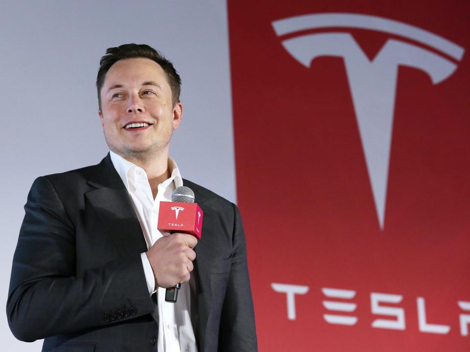 Elon Musk holding microphone in front of red sign with Tesla logo