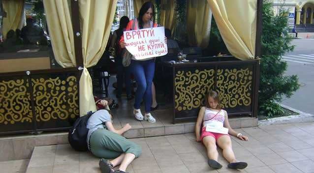 A man and a woman lie down outside a cafe, whilst another holds a sign up to the passing public. Photo: Facebook