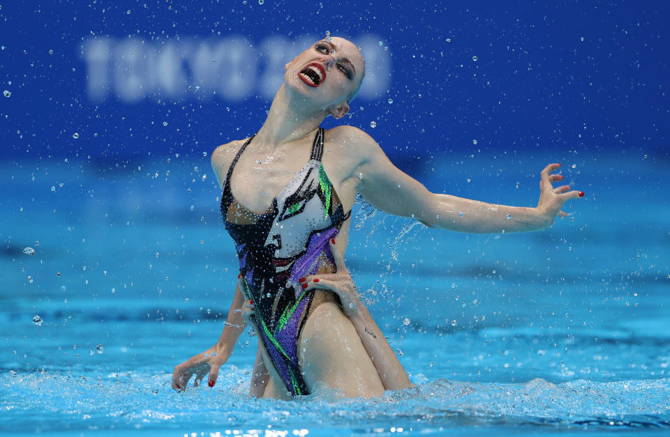 A synchronized swimmer being lifted out of the water during a routine with her mouth wide open and very red lips