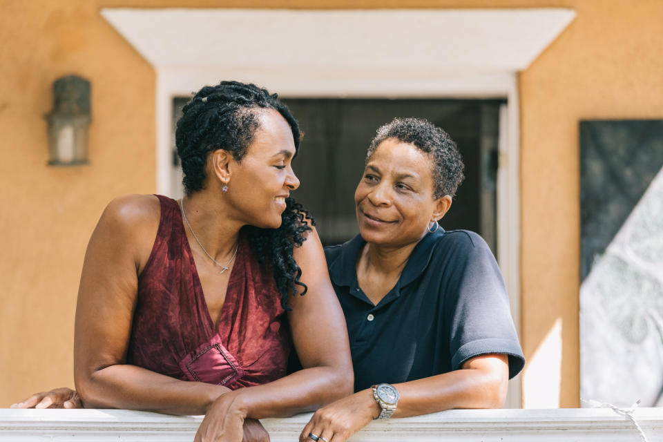 Two women are smiling at each other while leaning on a porch railing. Both appear happy and relaxed