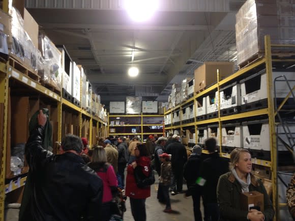 Aisle of warehouse store with pallets stacked to ceiling and dozens of customers shopping.