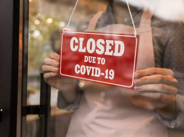 A woman puts a red sign with words Closed Due To COVID-19 onto a glass door. (Shutterstock - image credit)