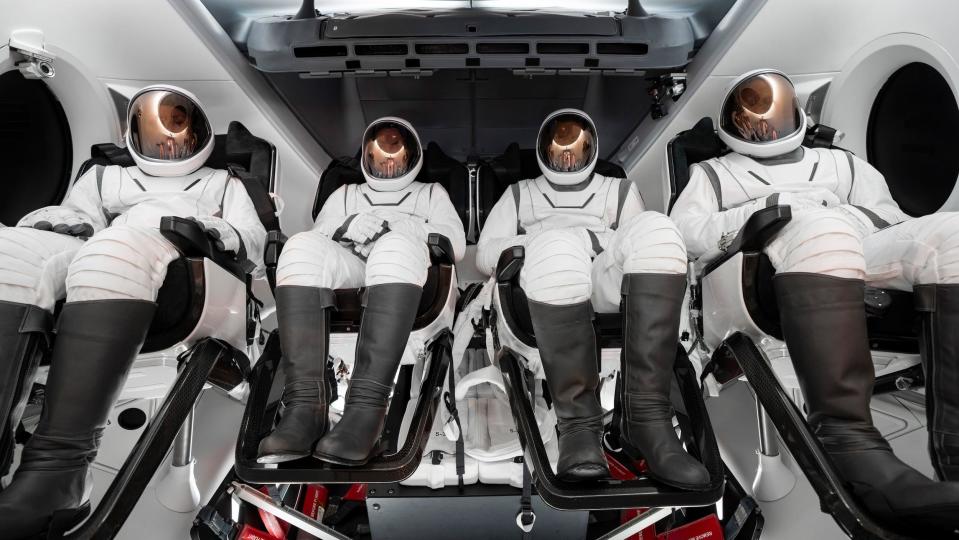 four people in white and gray spacesuits sit in a row of chairs with footrests in a spaceship