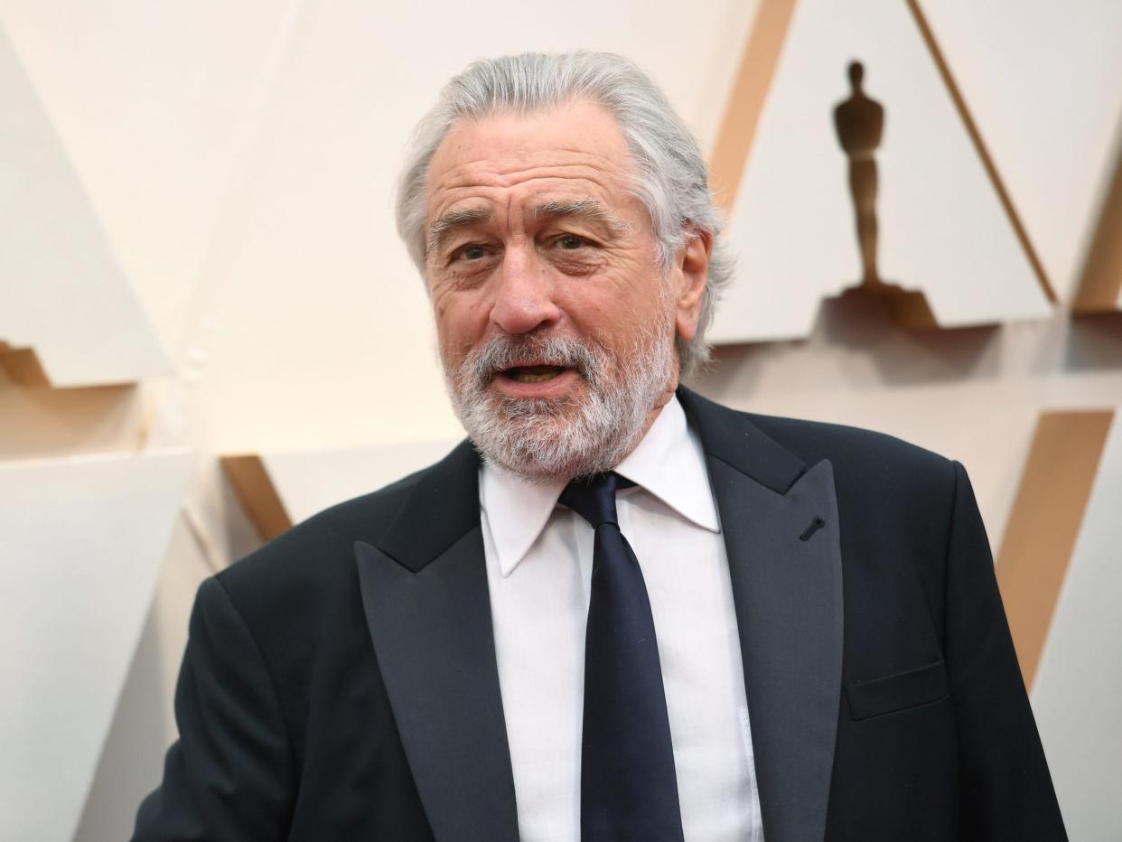 De Niro with grey hair and beard in a suit standing in front of an Oscars backdrop.