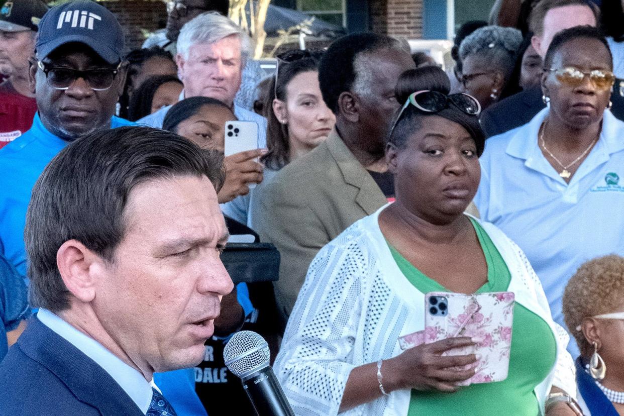 A white man speaks into a microphone. Standing beside him, a black woman looks skeptically on.