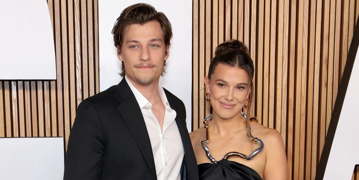 millie bobby brown wears a long black dress with a metallic necklace like neckline arm in arm with jake bongiovi who is dressed in a black suit and open neck white shirt