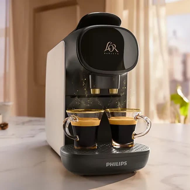 L'OR Barista Sublime coffee machine in white on a marble countertop in front of kitchen window. 