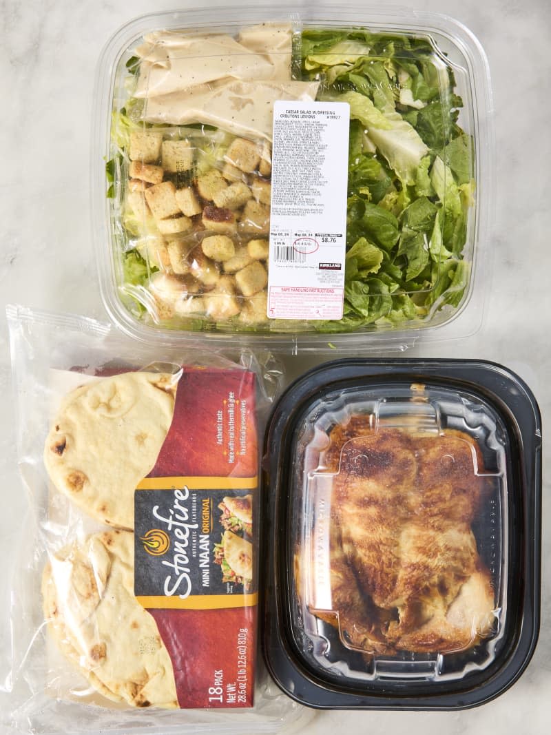 Costco ingredients to make a chicken caesar salad wrap laid out on a marble surface: naan, rotisserie chicken, and caesar salad