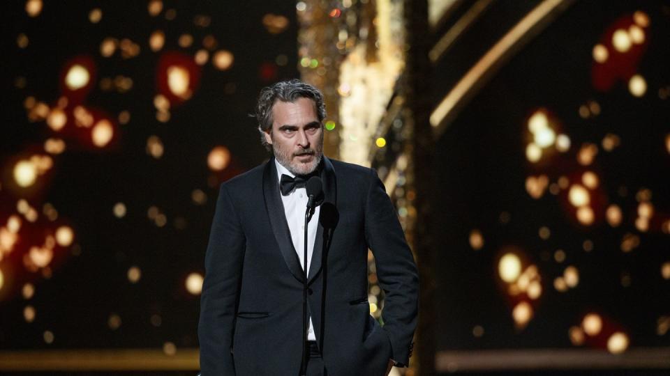 joaquin phoenix speaks into a microphone while standing on a stage, he wears a black suit with a black bowtie and white collared shirt, one hand holds an oscar statuette, his other hand is in his pocket