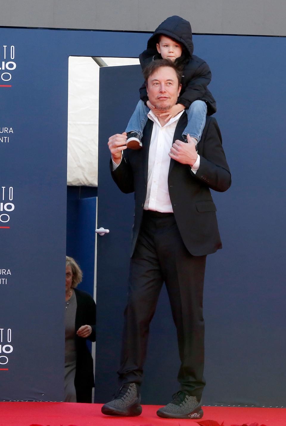 Elon Musk, wearing black shoes that say Cybertruck, and holding his son on his shoulders