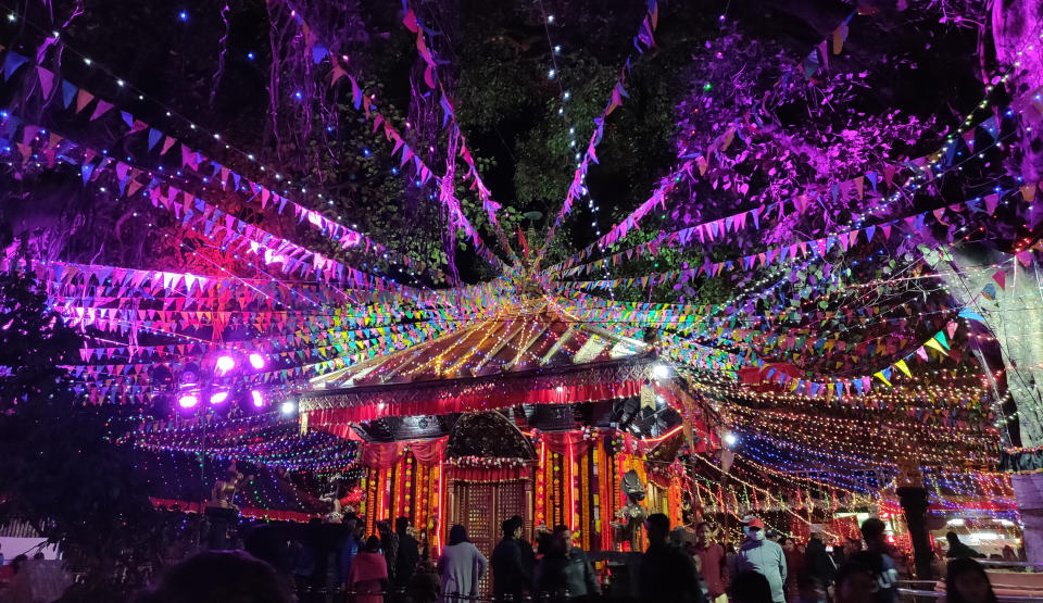 Outdoor festival at night under a canopy of colorful string lights and bunting, with a decorated pavilion in the center surrounded by people