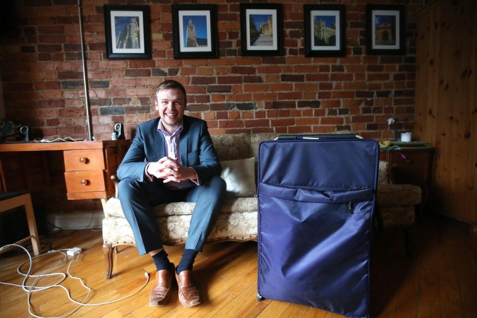 A man wearing a blue suit sitting on a white couch beside a desk in front of an exposed brick wall with a big blue suitcase next to him. He's in an Airbnb.