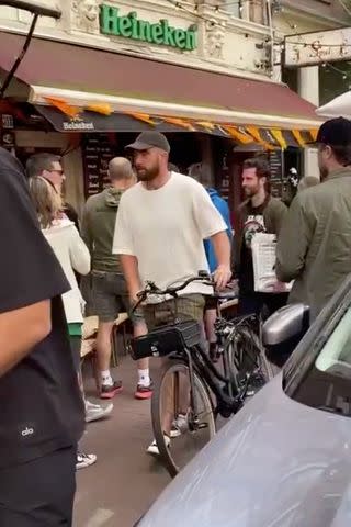 <p>Mark Gray for PEOPLE</p> Travis Kelce walks his bike on a busy street in Amsterdam on July 6, 2024