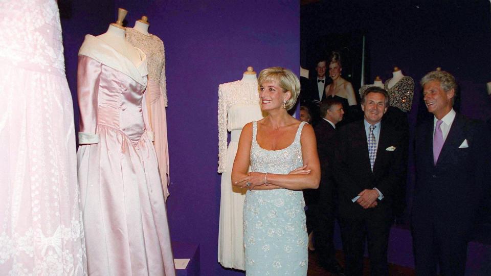 princess diana smiles as she looks at three dresses on mannequins, she wears a sleeveless minidress and clutches her elbows