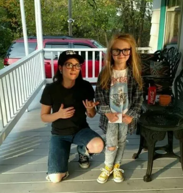 Person with glasses and wearing a T-shirt and baseball cap next to a kid with long blonde hair and glasses and wearing a flannel shirt and jeans